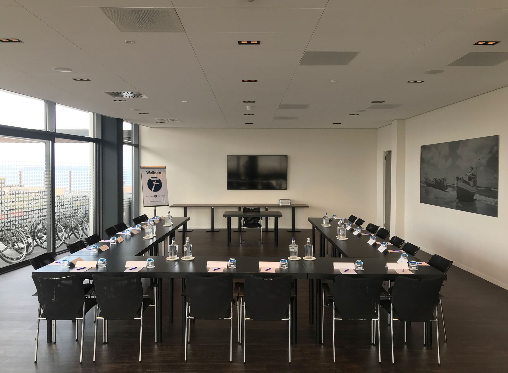 A conference room with a long table, chairs, name cards, water bottles, and a city view through large windows.