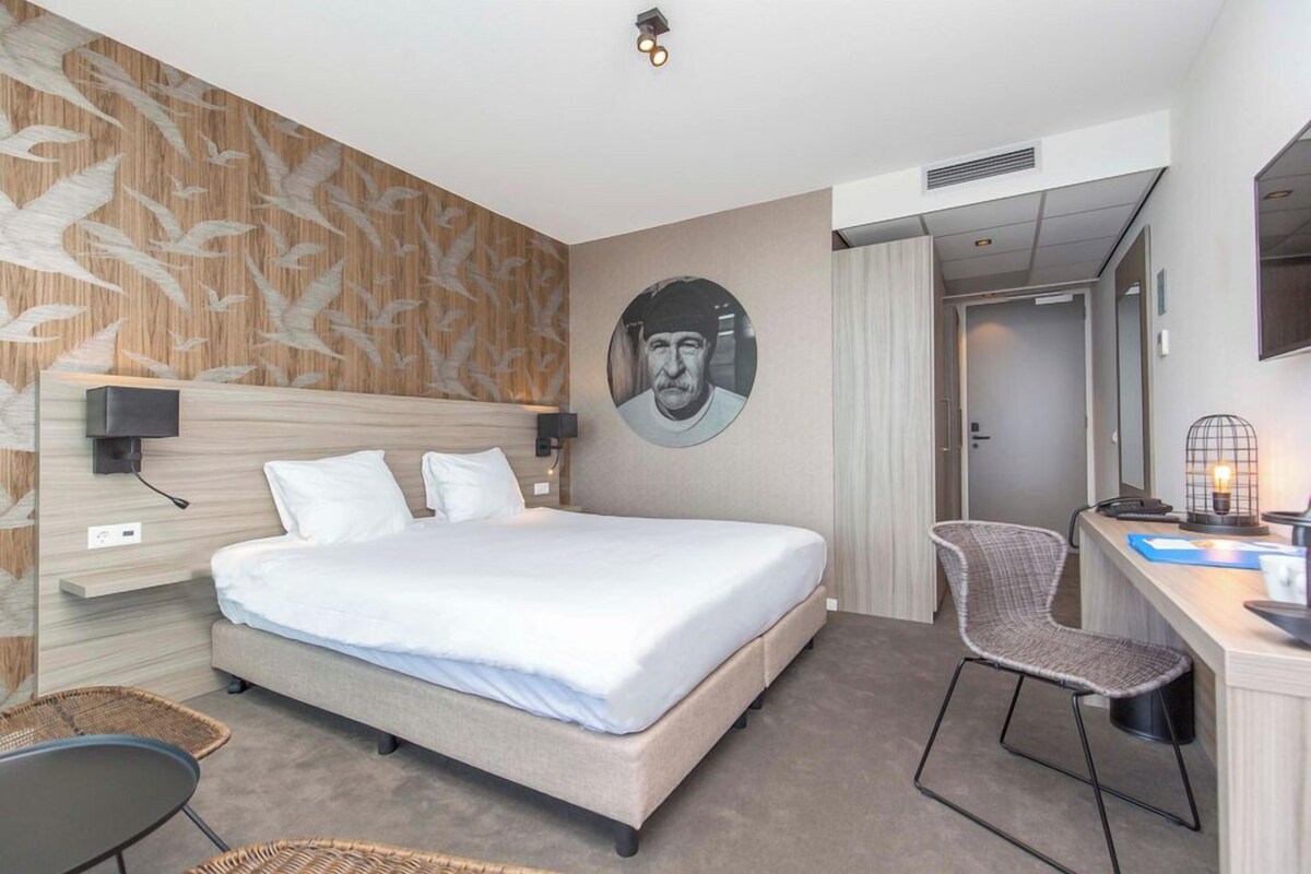 A hotel room with a bed, a desk with a chair, a wall-mounted TV, and a circular portrait.