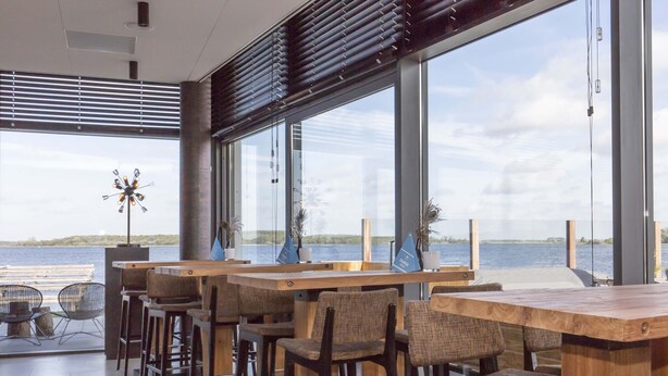 Un espace bar moderne avec des tables et des chaises en bois, de grandes fenêtres offrant une vue sur l’eau et un luminaire décoratif.