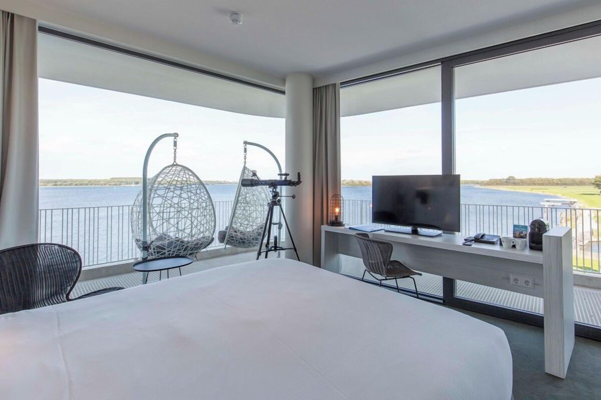 A hotel room with a large bed, a desk with a TV, two hanging chairs, and a view of the water.