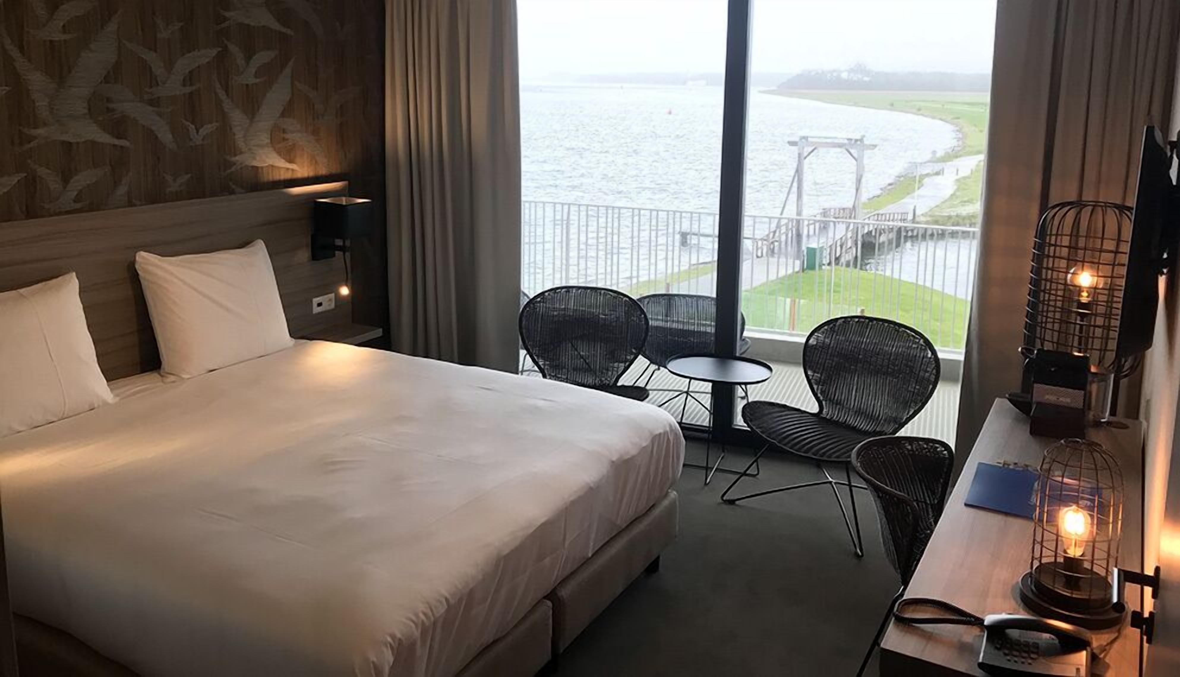 Une chambre d’hôtel avec un grand lit, deux fauteuils, une petite table et une vue sur une rivière et un pont.