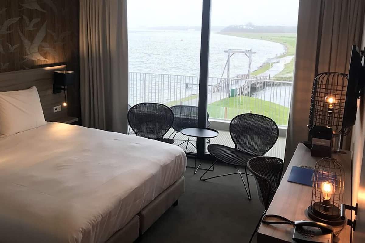 Ein Hotelzimmer mit einem großen Bett, zwei Stühlen, einem kleinen Tisch und Blick auf einen Fluss und eine Brücke.
