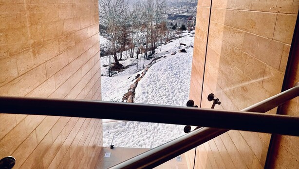 Vista desde una ventana de madera que da a un paisaje nevado con árboles desnudos y edificios a la distancia.