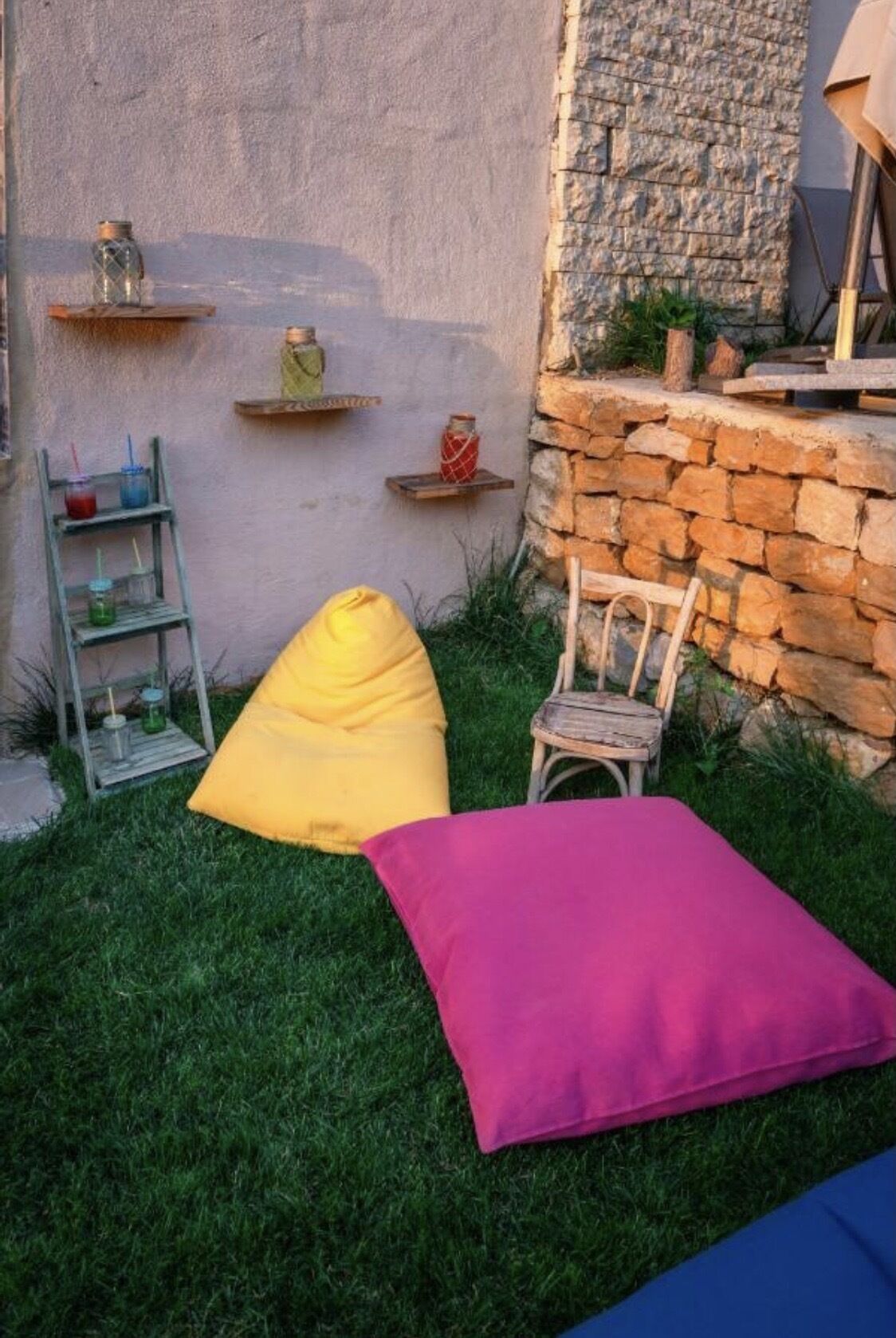 Un pequeño espacio al aire libre con un sillón de mezcla amarilla y un cojín rosa sobre un césped, un rústico muro de piedra y una estantería metálica con bebidas.