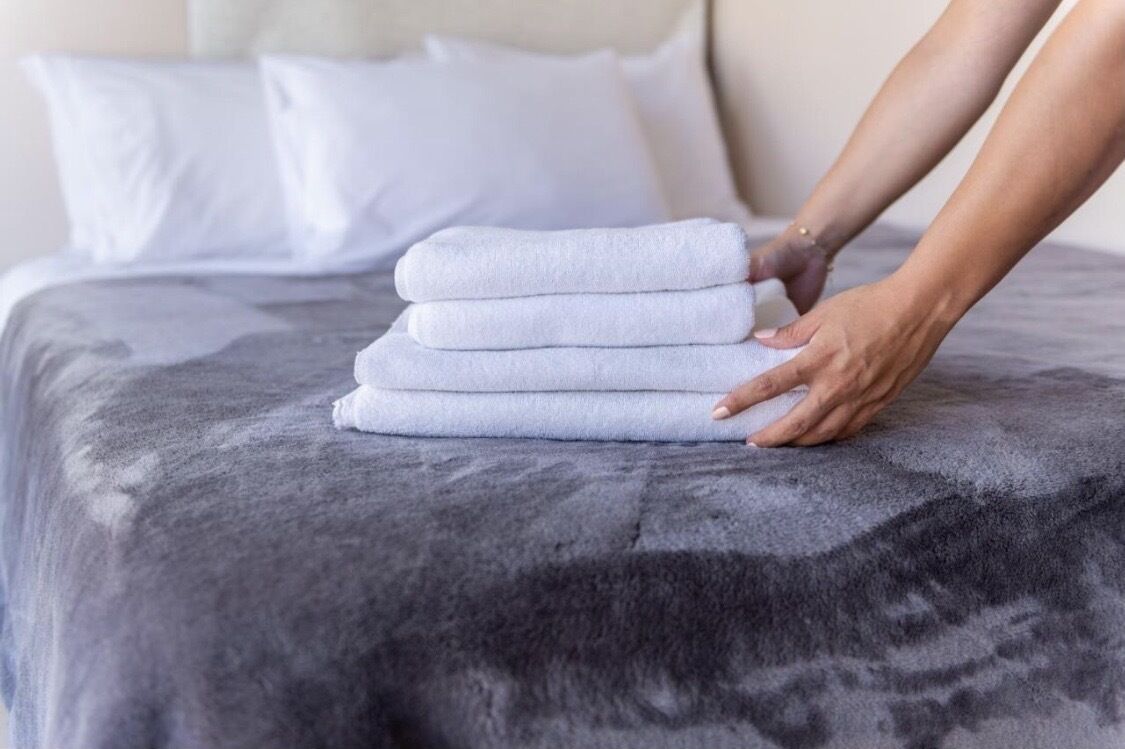 Un lit bien fait, recouvert d’une couverture grise et sur lequel est posée une pile de serviettes blanches.