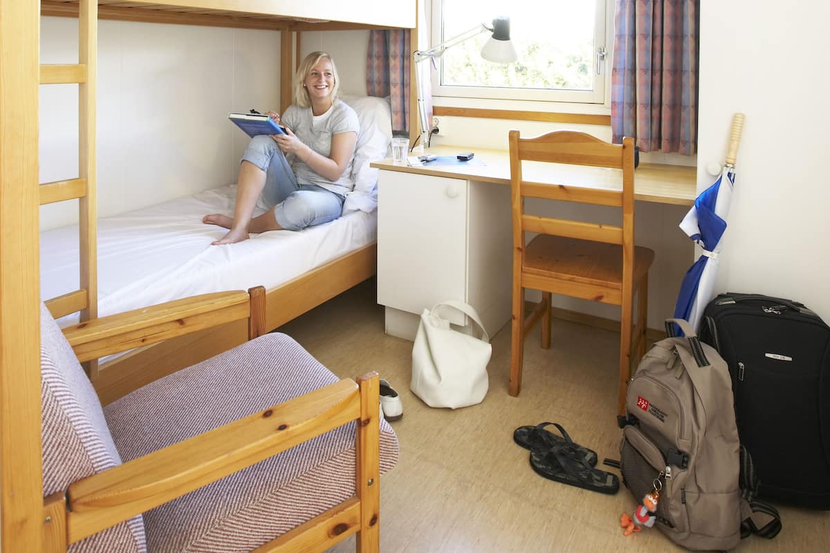 Ein Schlafsaalzimmer mit Etagenbetten, einem Schreibtisch, einem Stuhl, einem Fenster mit Vorhängen und einem Rucksack auf dem Boden.