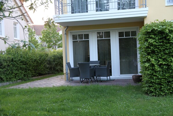 Un patio con mesa y sillas, rodeado de follaje y un edificio con balcón.