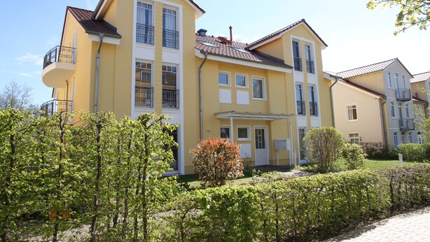 A multi-story yellow building with balconies, a covered entrance, and a well-maintained garden. - Ferienoase Usedom (Heringsdorf)