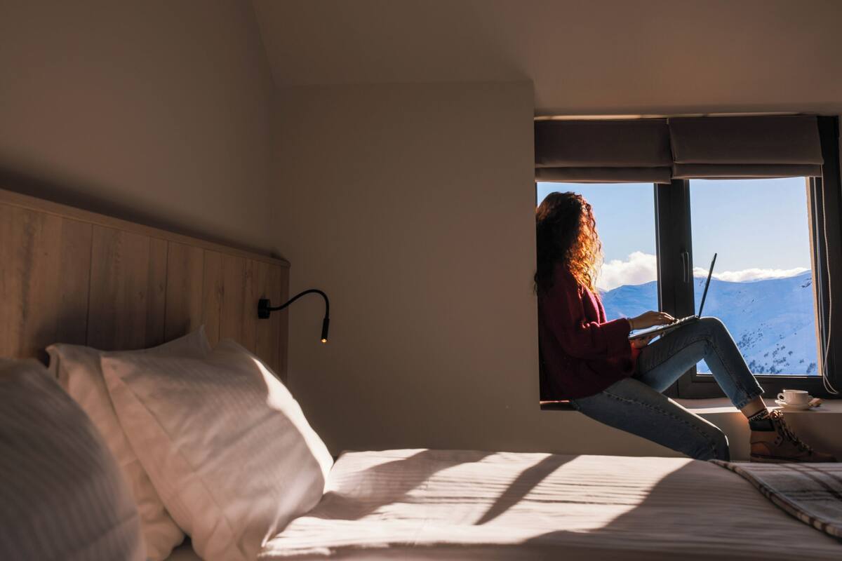 A person sitting on a bed with a laptop, using a window with a view of mountains.