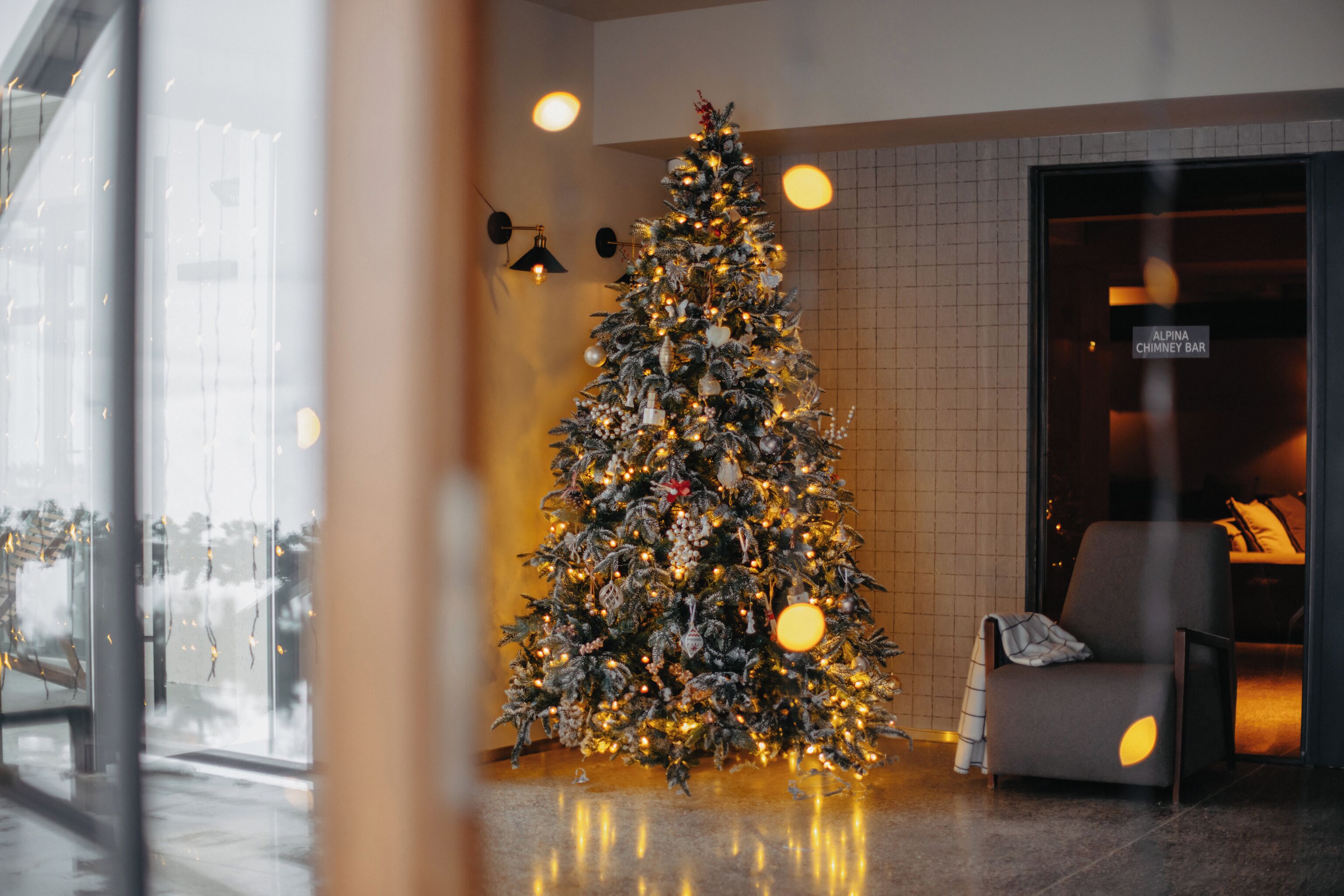 A Christmas tree decorated with lights and ornaments in a hotel lobby with a modern design, a seating area, and a sign for the Alpine Chimney Bar.