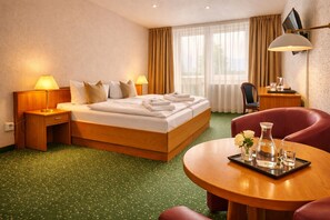 A hotel room with a bed, two bedside tables with lamps, a desk with a chair, a round table with a vase and glasses, and a TV mounted on the wall. - Lázeňský Hotel Pyramida I (Frantiskovy Lazne)