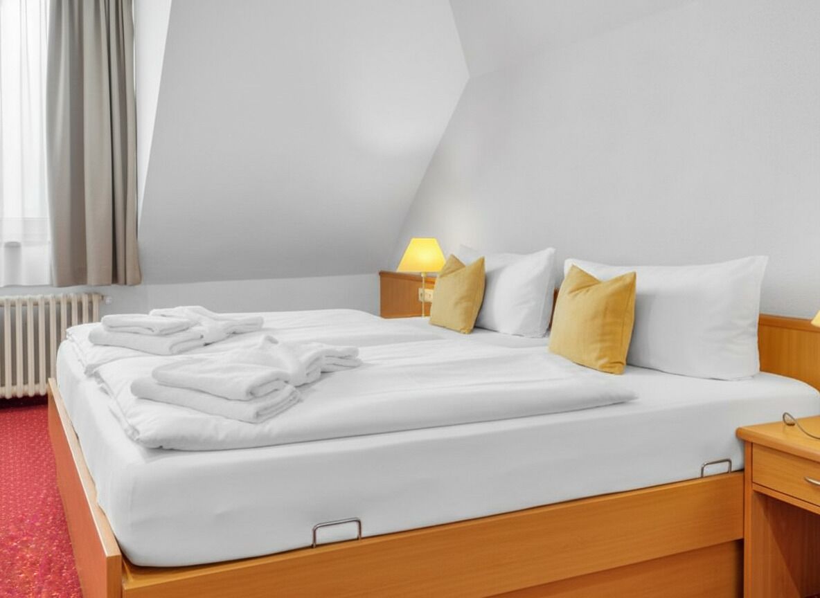 A neatly made bed with white linens and a folded towel, a bedside lamp, and a nightstand with a lamp.