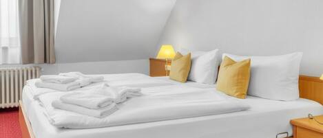 A neatly made bed with white linens and a folded towel, a bedside lamp, and a nightstand with a lamp.