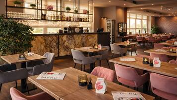 Un restaurante con mesas de madera, sillas acolchadas, una barra de mármol y estantes con cristalería y plantas.