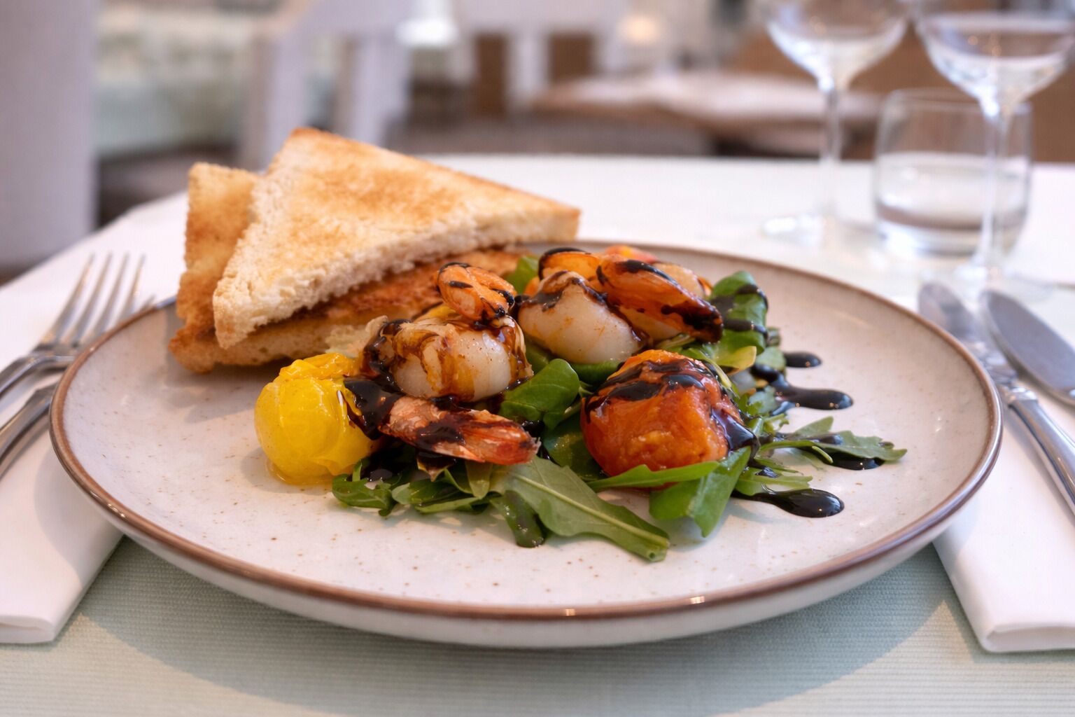 A plate of food with grilled shrimp, lemon, and greens, accompanied by a slice of bread on a table.
