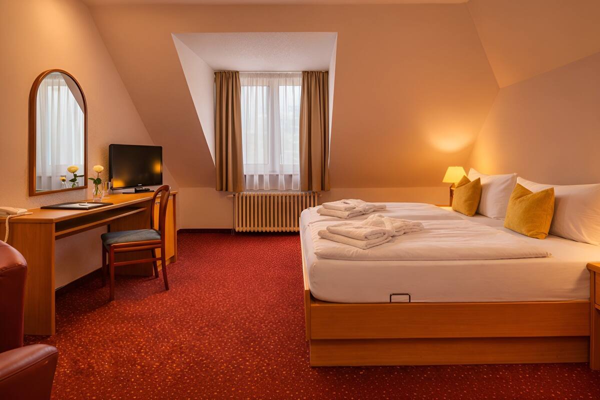 A hotel room with a bed, a desk with a chair, a television, and a window with curtains.