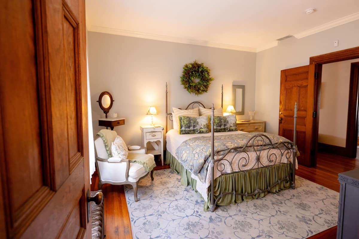 A bedroom with a four-poster bed, a nightstand, a chair, a mirror, and a wall-mounted wreath.