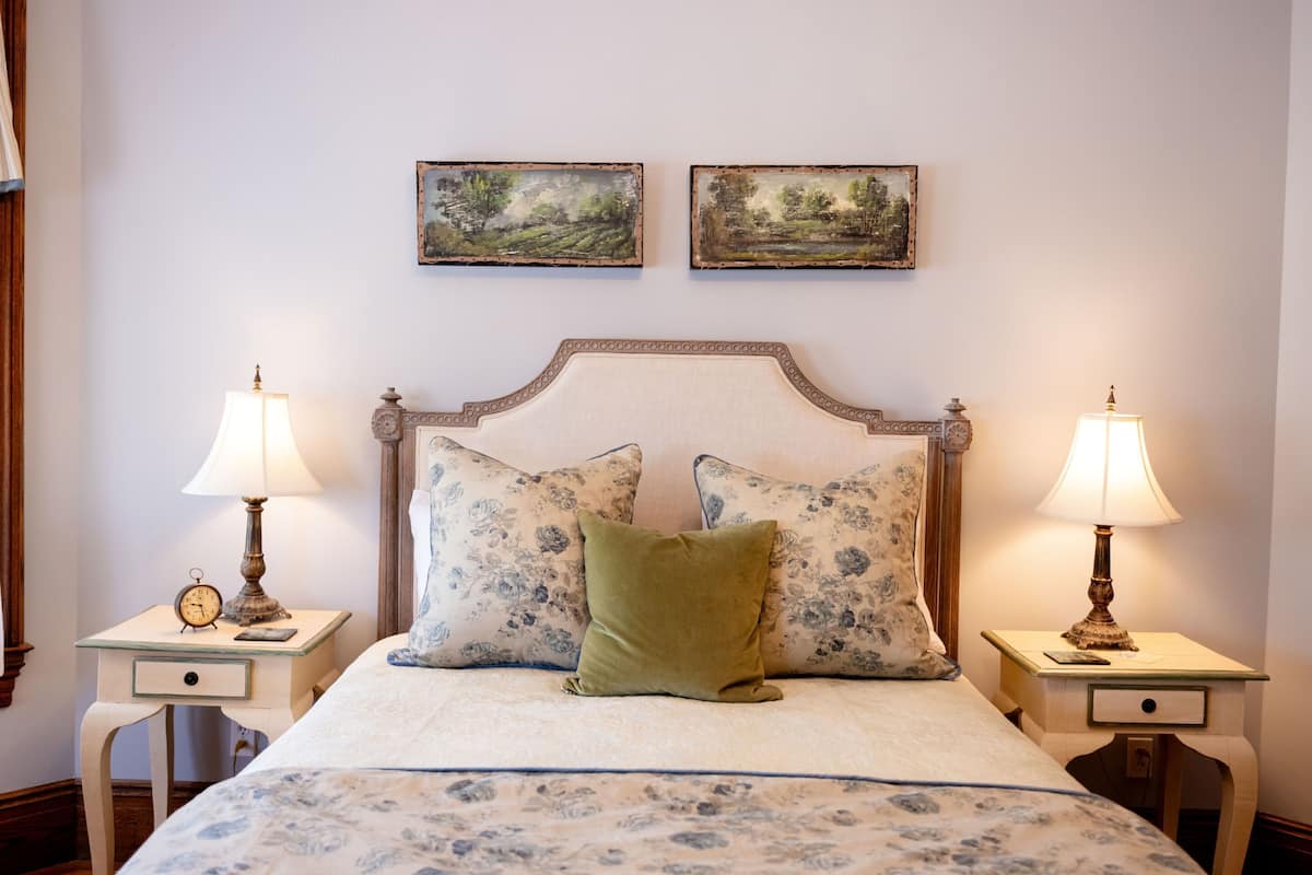 A bed with a floral-patterned bedspread, two matching nightstands with lamps, and two framed landscape paintings on the wall.