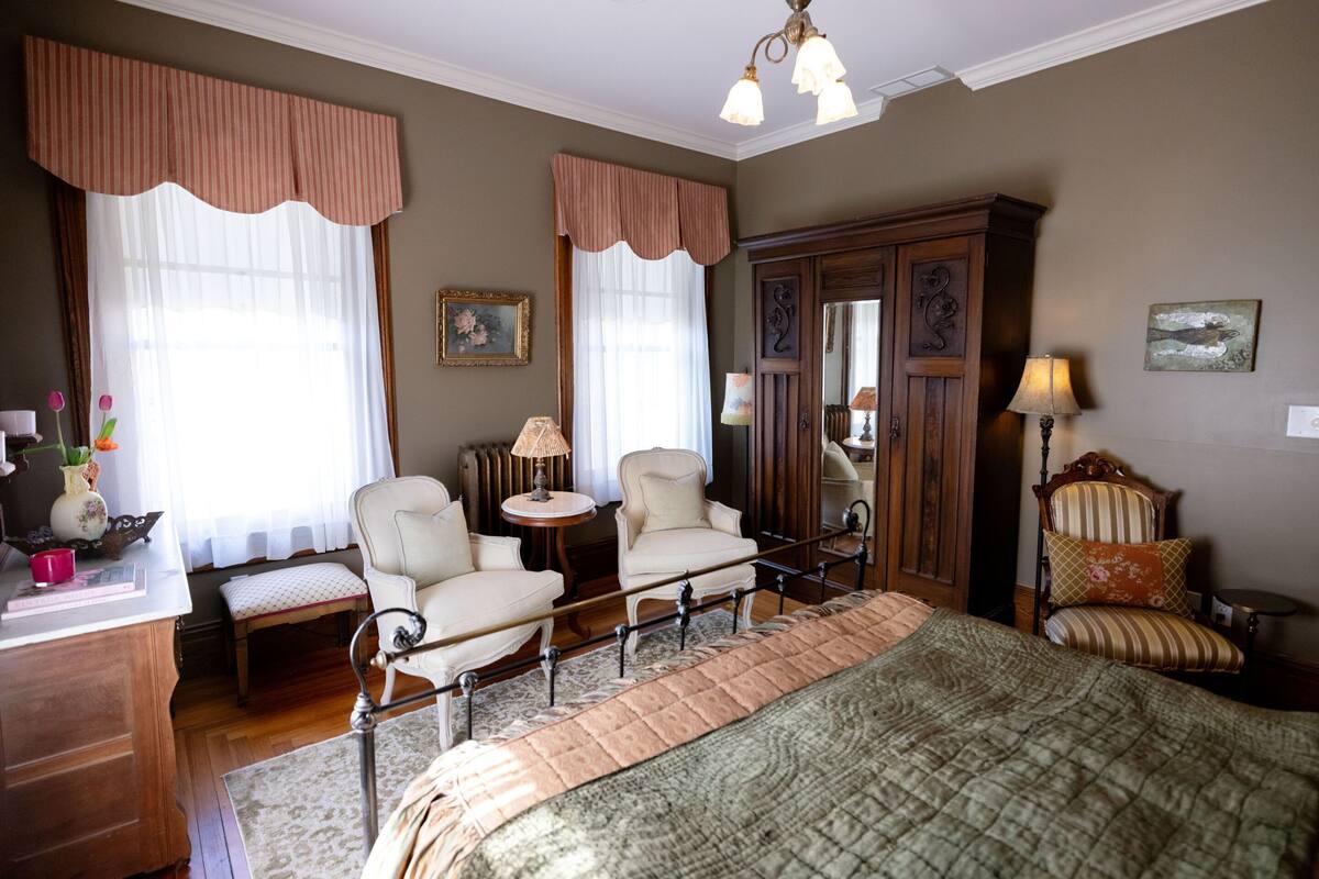 A bedroom with a bed, two chairs, a nightstand, a dresser, and a mirror.