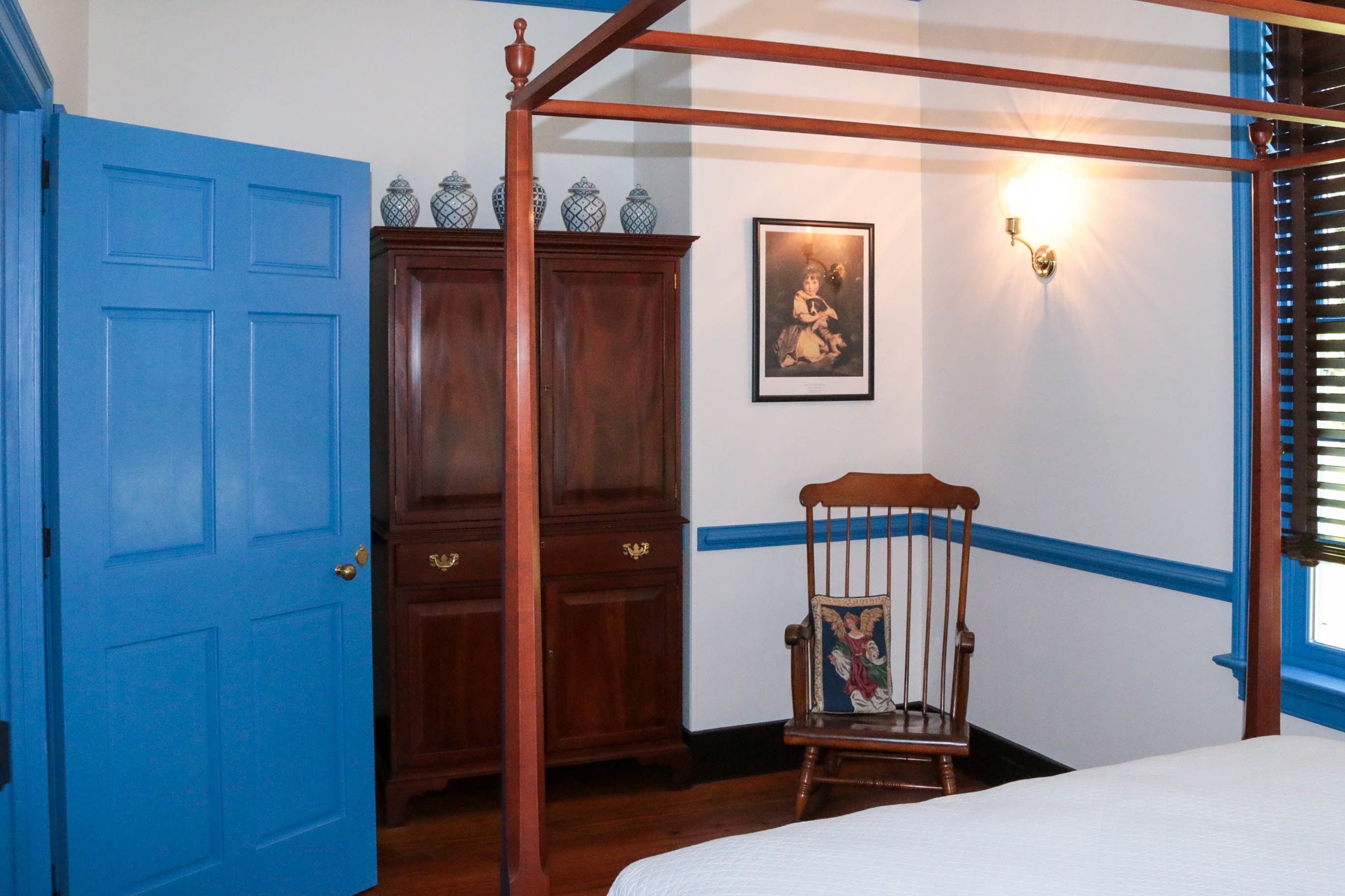 A bedroom with a four-poster bed, a wooden wardrobe, a chair, and a wall-mounted light fixture.