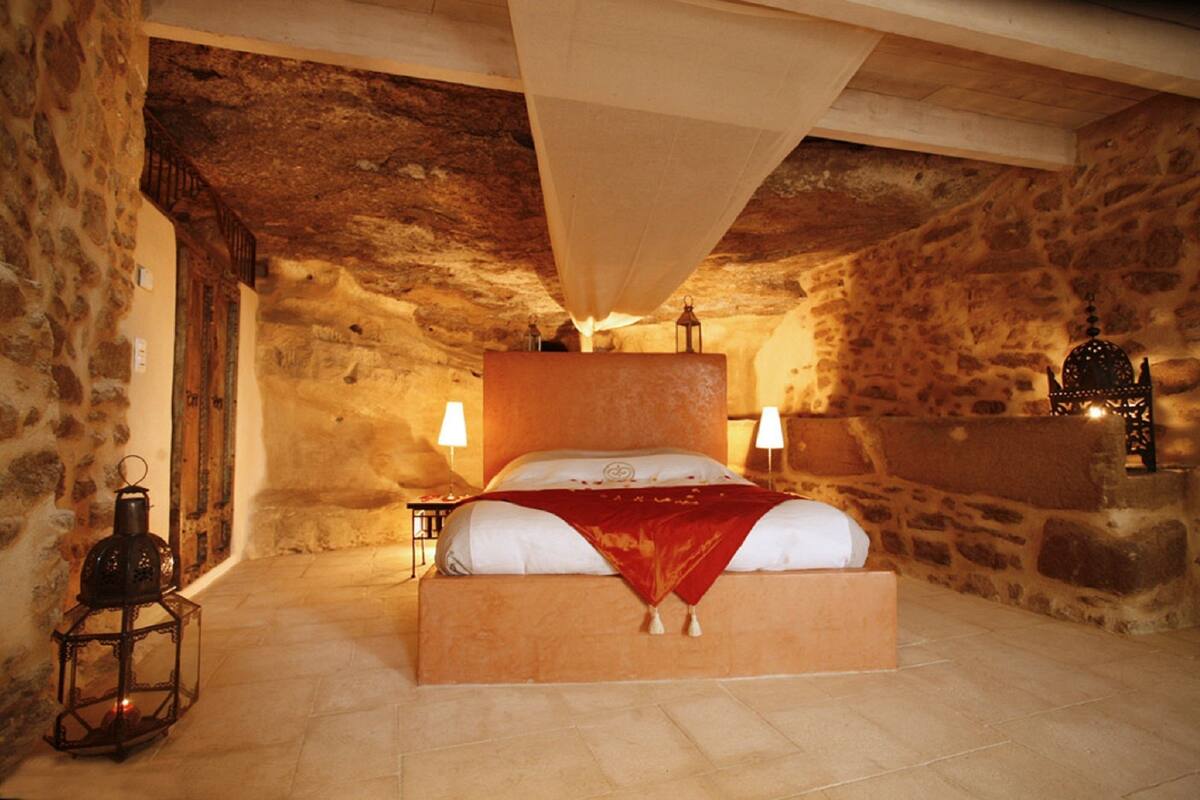 A stone-walled bedroom with a large bed, two bedside lamps, and a lantern on the floor.