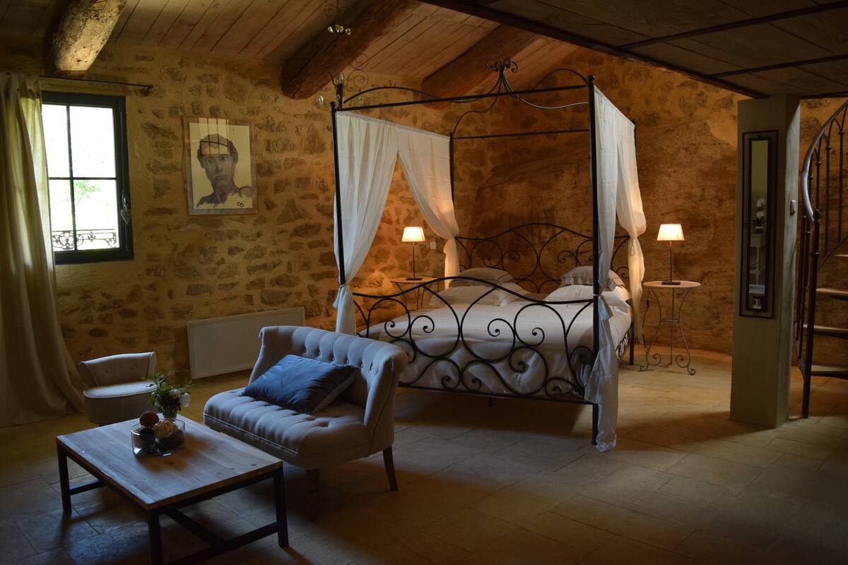 A bedroom with a four-poster bed, a sitting area with a table and chair, and a stone wall.