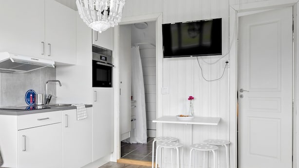 A modern kitchen with white cabinets, a built-in oven, a stainless steel countertop, and a TV mounted on the wall. There are two white stools and a small table with a vase. - First Camp Hagön Halmstad (Halmstad)