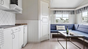 A compact kitchen with white cabinetry, a microwave, and a stove. A blue sofa with a cushion is positioned near a window with checkered curtains. - First Camp Hagön Halmstad (Halmstad)