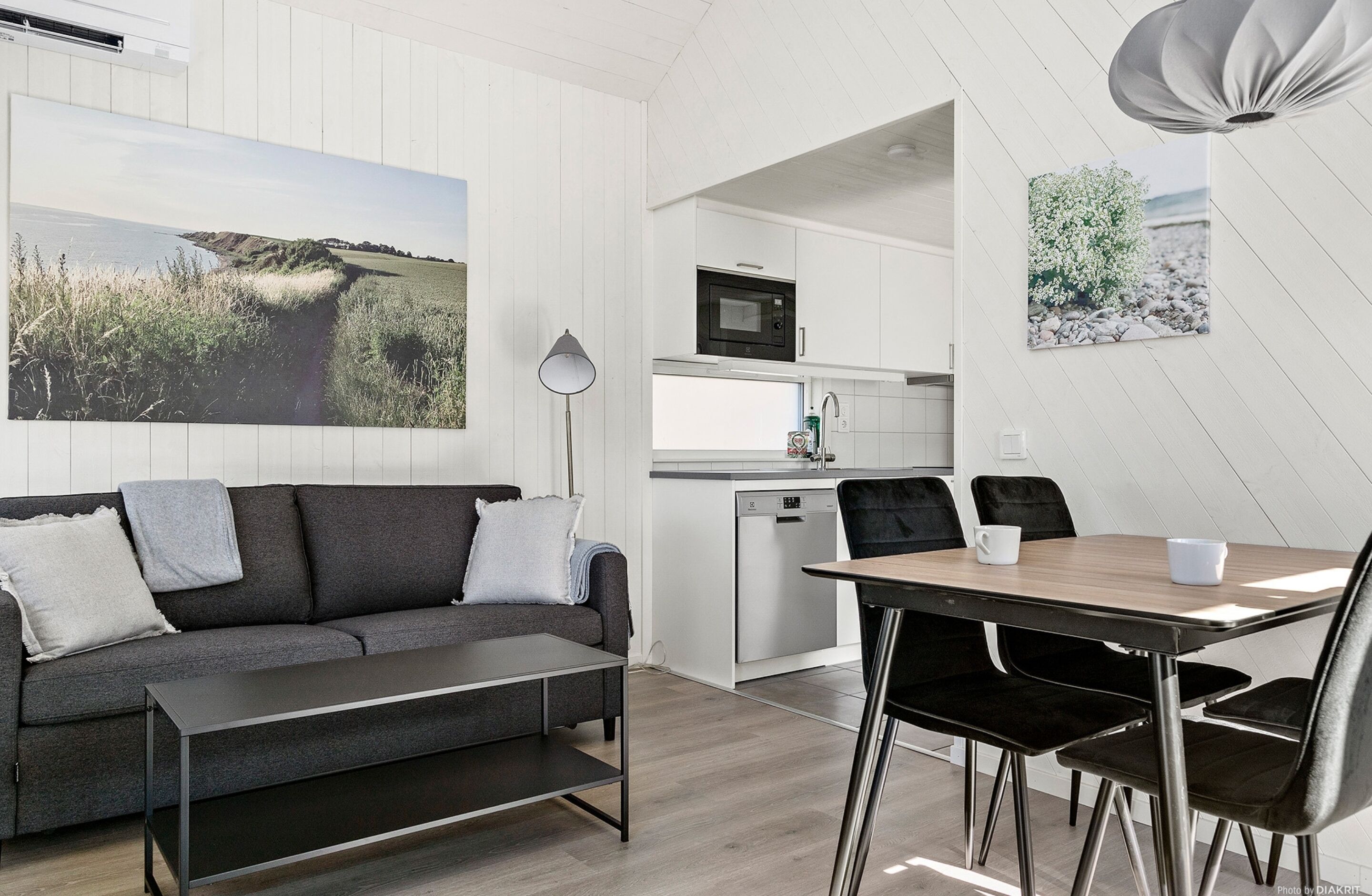 A modern living room with a sofa, a dining table with chairs, a kitchen area with a microwave, and a large landscape painting on the wall.