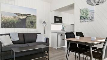 A modern living room with a sofa, a dining table with chairs, a kitchen area with a microwave, and a large landscape painting on the wall.