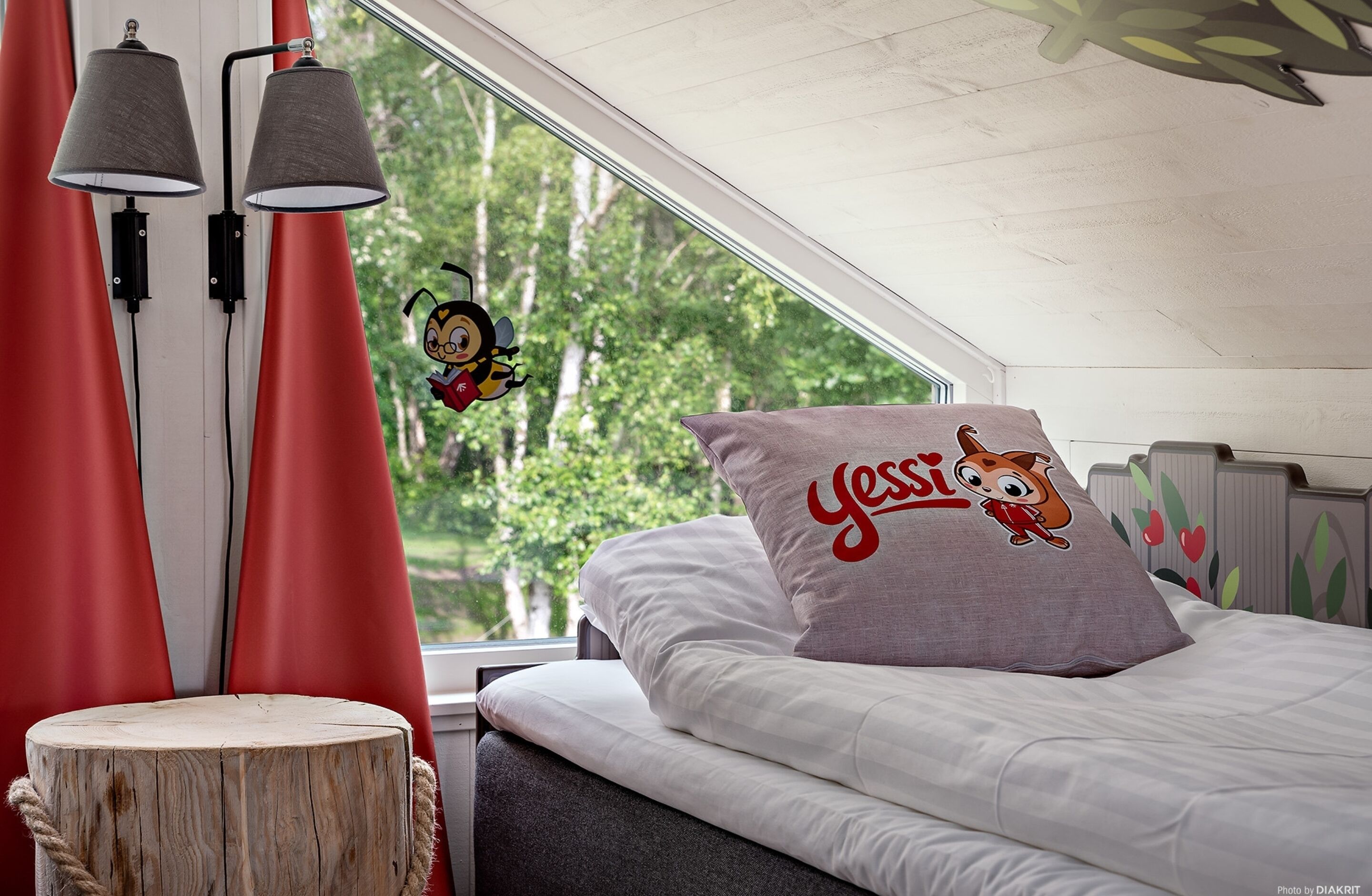 A bedroom with a large window, a bed with a pillow featuring a cartoon character, and a wooden stool.