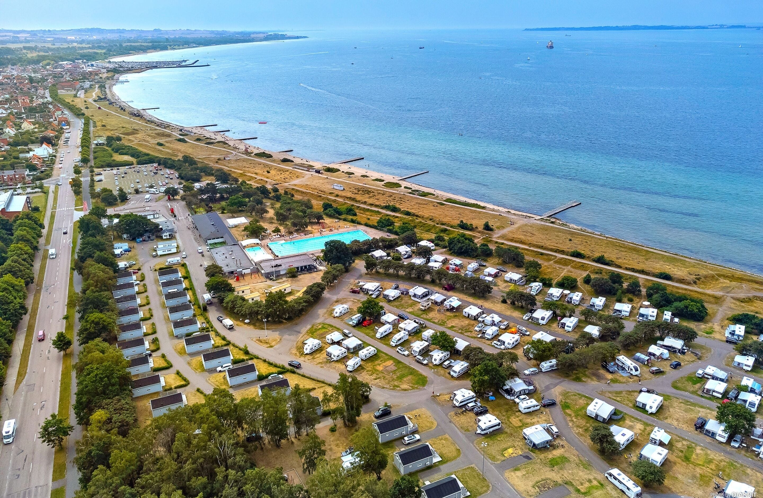 Vista aérea de un parque para vehículos recreativos en la costa, con varias autocaravanas, una piscina y una playa.