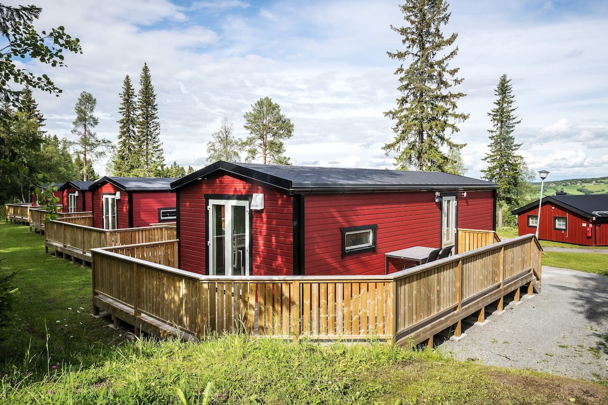A row of red cabins with wooden decks, surrounded by tall trees and a clear sky.