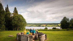 Un grupo de personas alrededor de un brasero de madera en un campo con césped, con un lago y árboles al fondo.