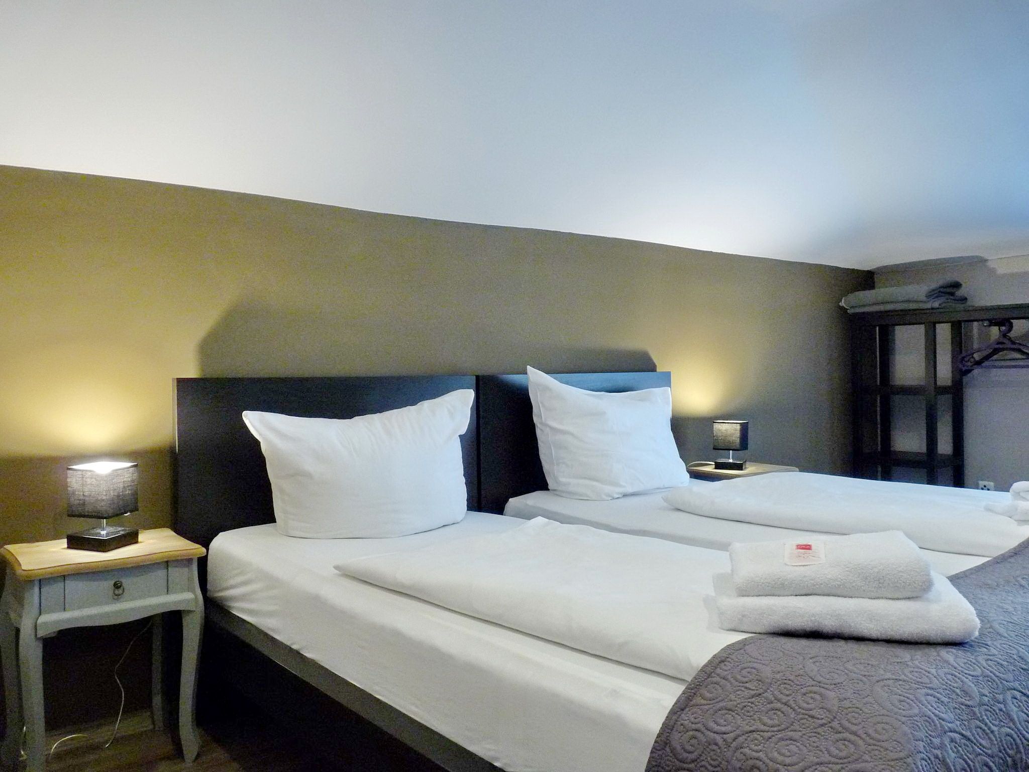 Habitación de hotel con dos camas, cada una con ropa blanca y almohadas, una mesita de noche con lámpara, y un armario con espejo.