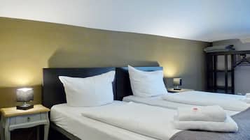 Habitación de hotel con dos camas, cada una con ropa blanca y almohadas, una mesita de noche con lámpara, y un armario con espejo.