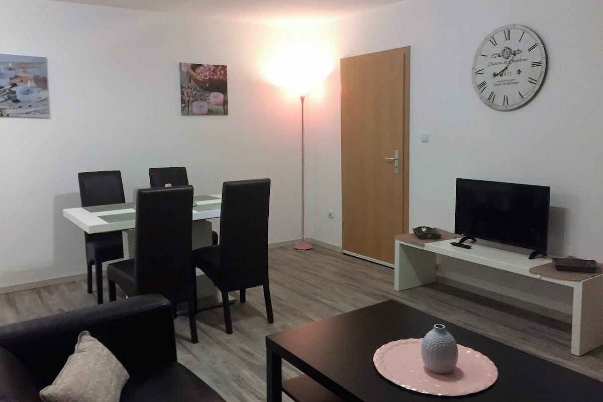 A modern living room with a dining area, a flat-screen TV, and a wall clock.