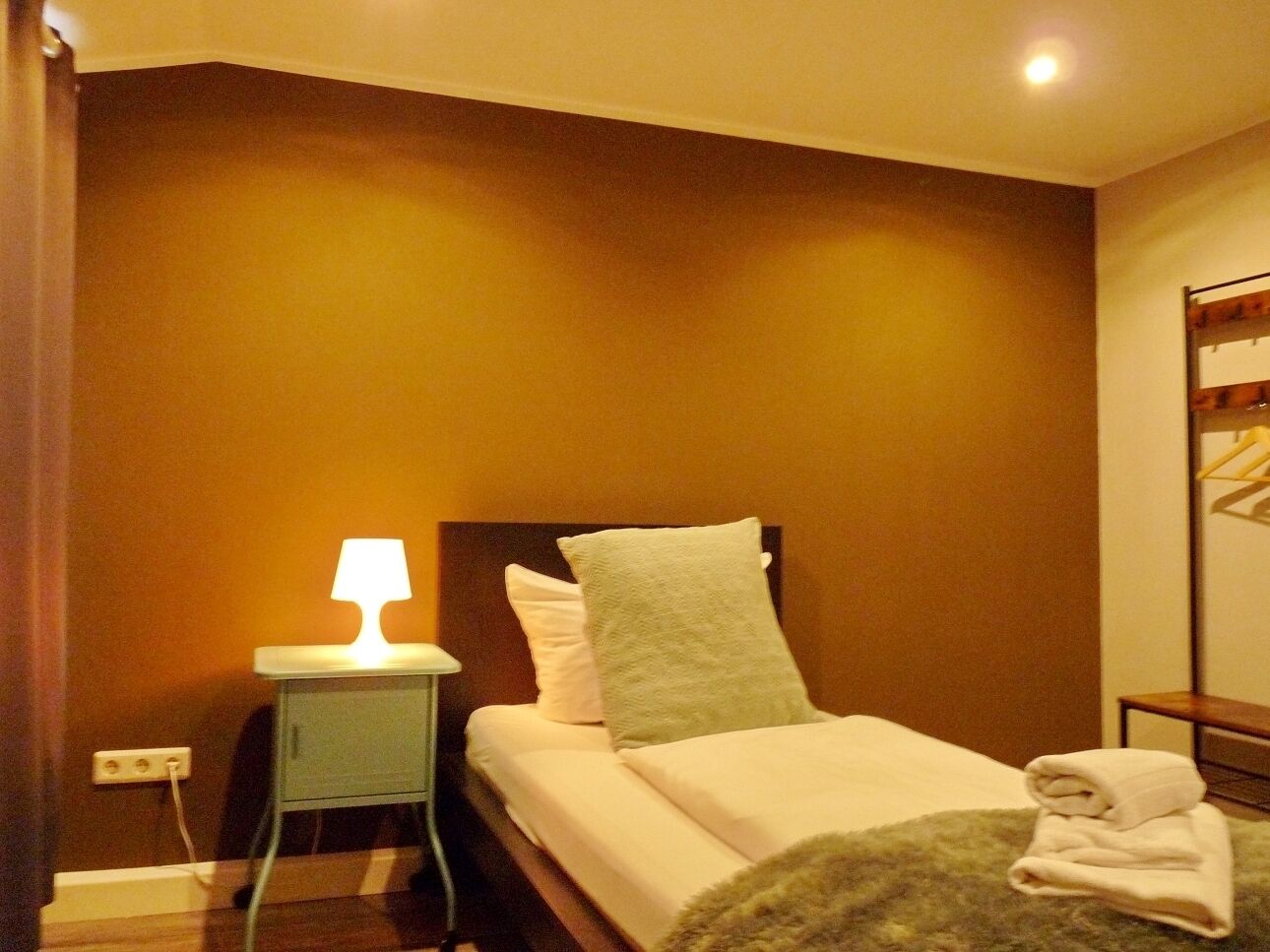 A hotel room with a bed, a bedside table with a lamp, and a wall-mounted coat rack.