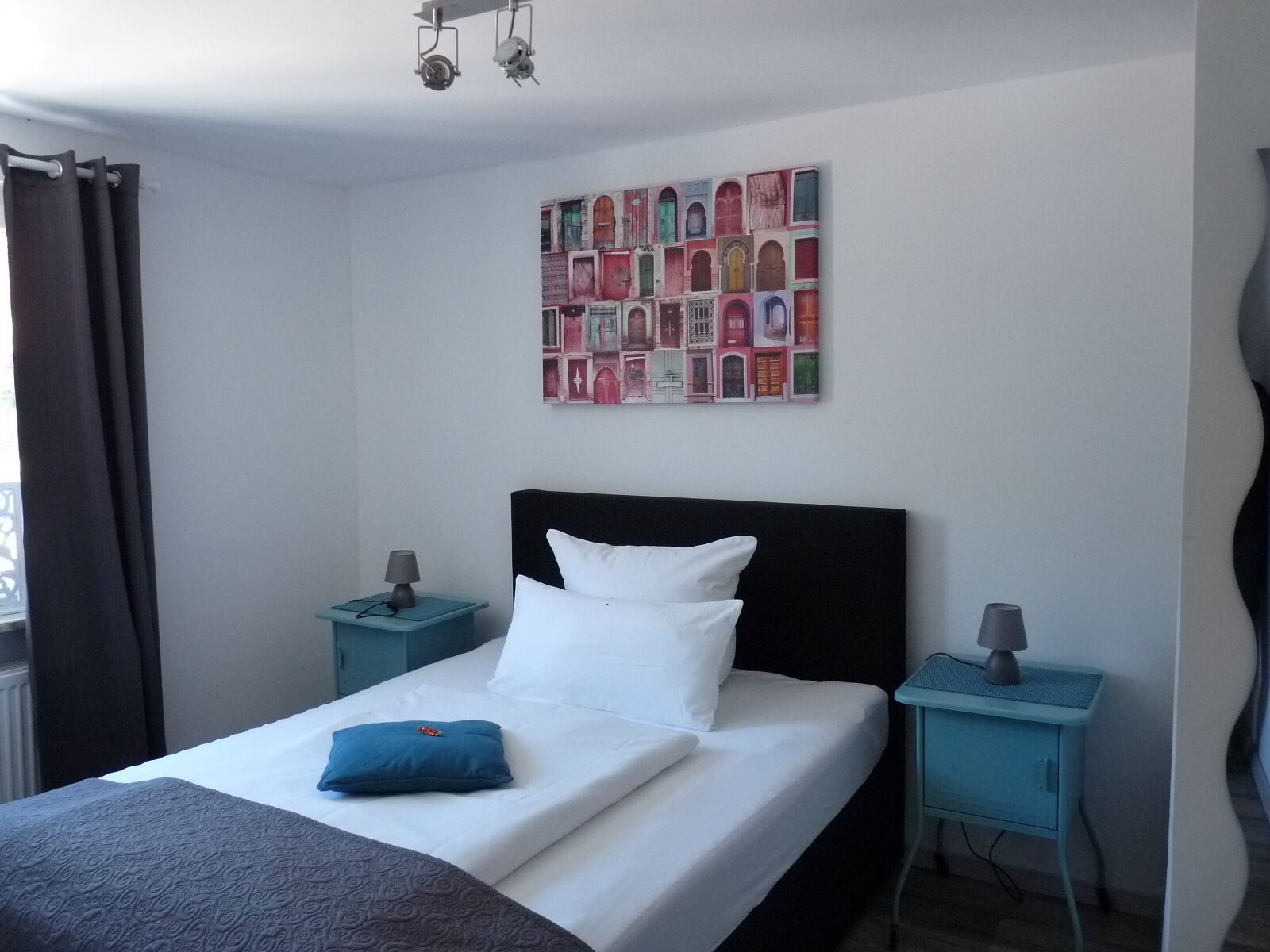 Een net opgemaakt bed met wit en blauw beddengoed, twee nachtkastjes met lampen, een kunstwerk aan de muur en een raam met gordijnen.
