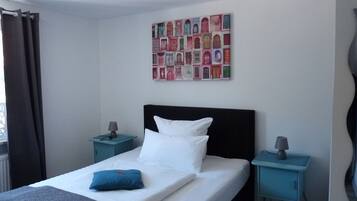 Una cama bien hecha con ropa de cama blanca y azul, dos mesitas de noche con lámparas, una obra de arte en la pared y una ventana con cortinas.