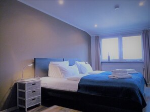A hotel room with a bed, a nightstand, a lamp, a window with curtains, and a view of the city. - Haus am Burggraben (Hinte)