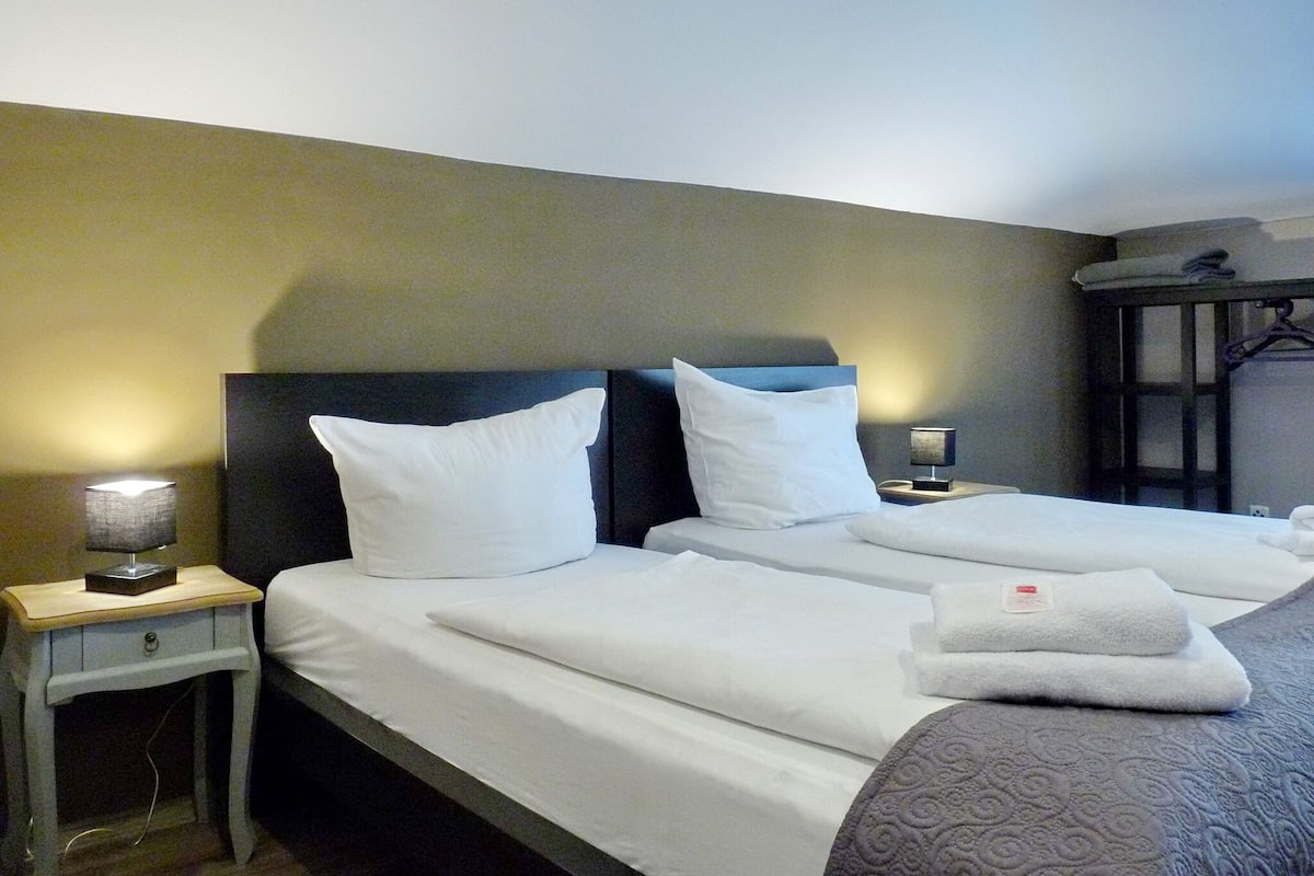 A hotel room with two beds, each with white linens and pillows, a bedside table with a lamp, and a wardrobe with a mirror.