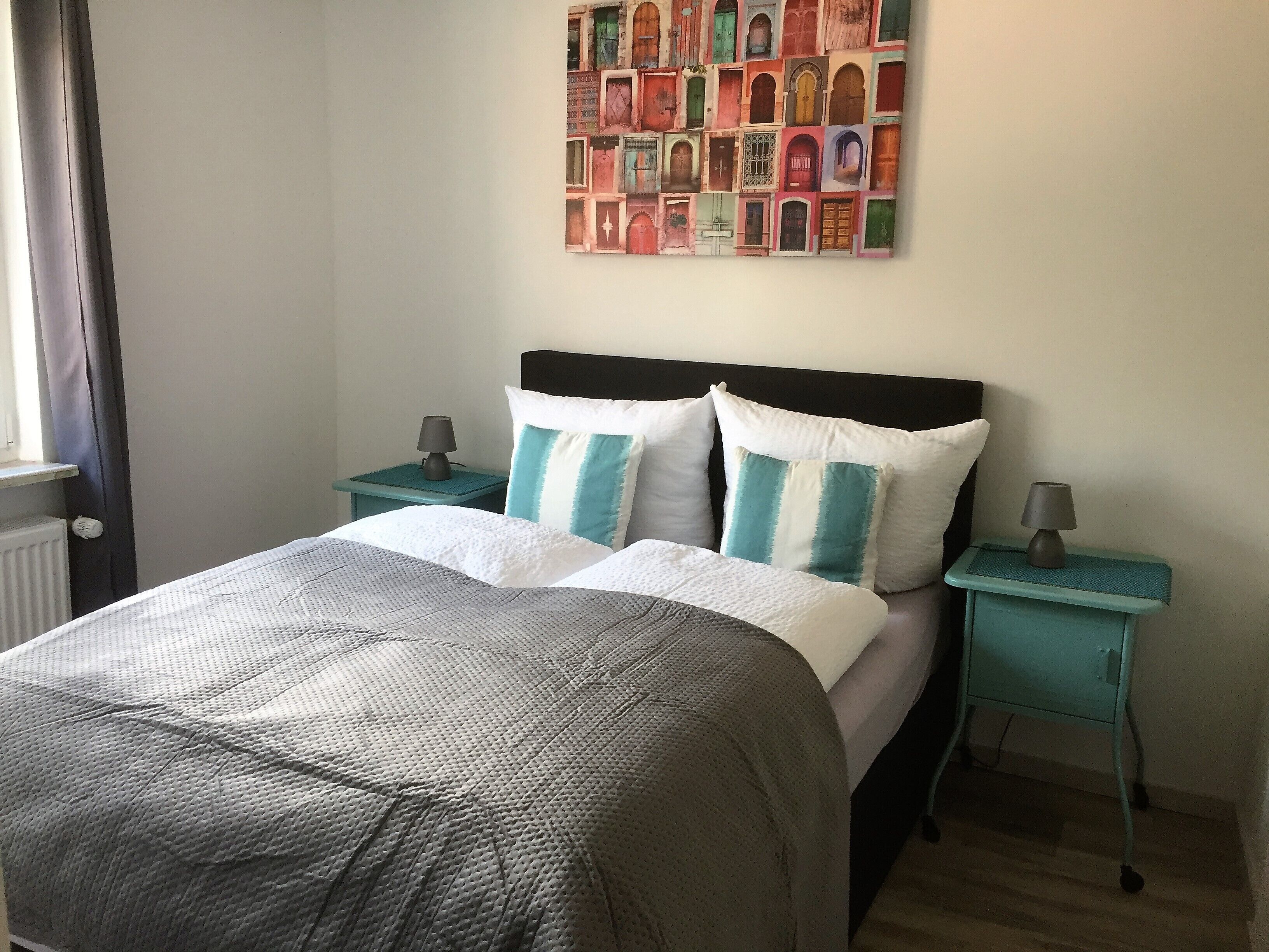 A neatly made bed with a grey and white duvet, two matching pillows, and a headboard. There are two bedside tables with lamps and a wall art piece above the bed.