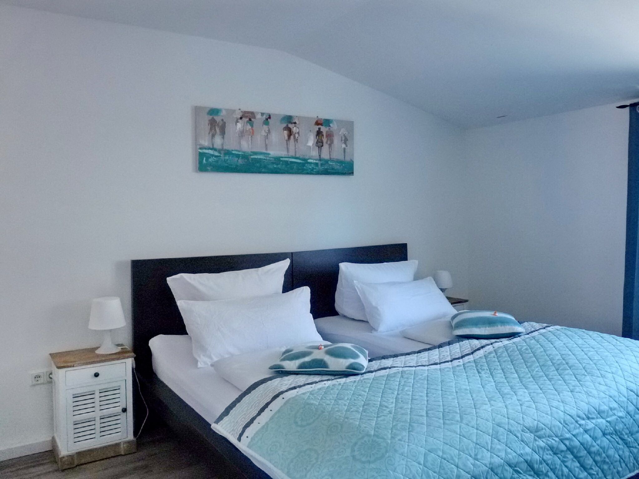Ein ordentlich bezogenes Bett mit weißer und blauer Bettwäsche, ein kleiner Nachttisch mit Lampe und ein Wandbild über dem Bett.