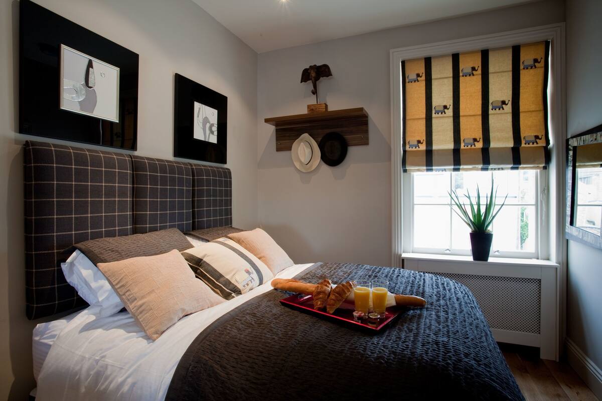 A neatly made bed with a tray of croissants and juice, a clock, and a window with a plant.