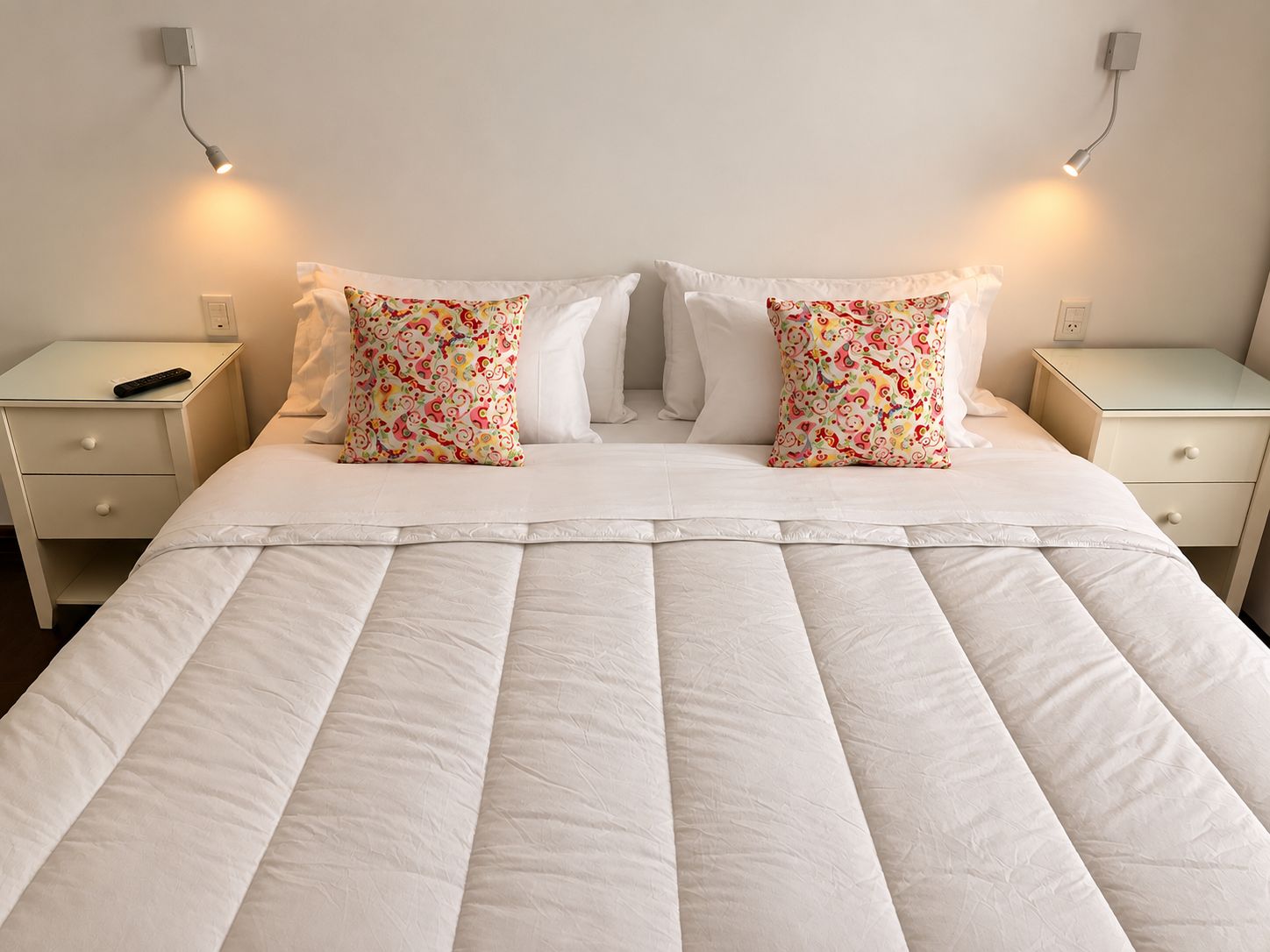 A neatly made bed with white bedding and two decorative pillows, flanked by two bedside tables with a remote control on one.