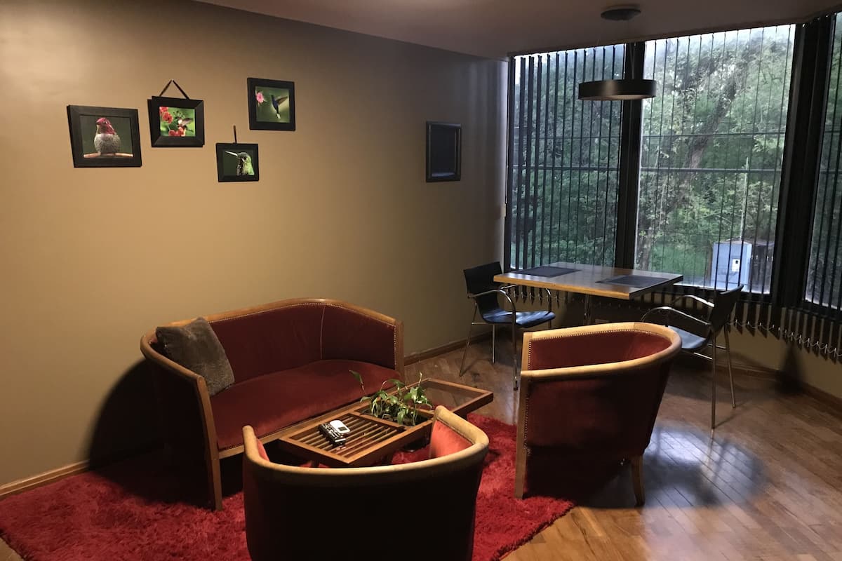 Un salon avec un canapé rouge, une table basse et une table à manger avec des chaises. Des tableaux encadrés ornent le mur, et une fenêtre offre une vue sur les arbres.