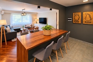 Una sala de estar moderna con una mesa de comedor de madera, sillas tapizadas en gris, un sofá, un televisor y un ventanal con cortinas.