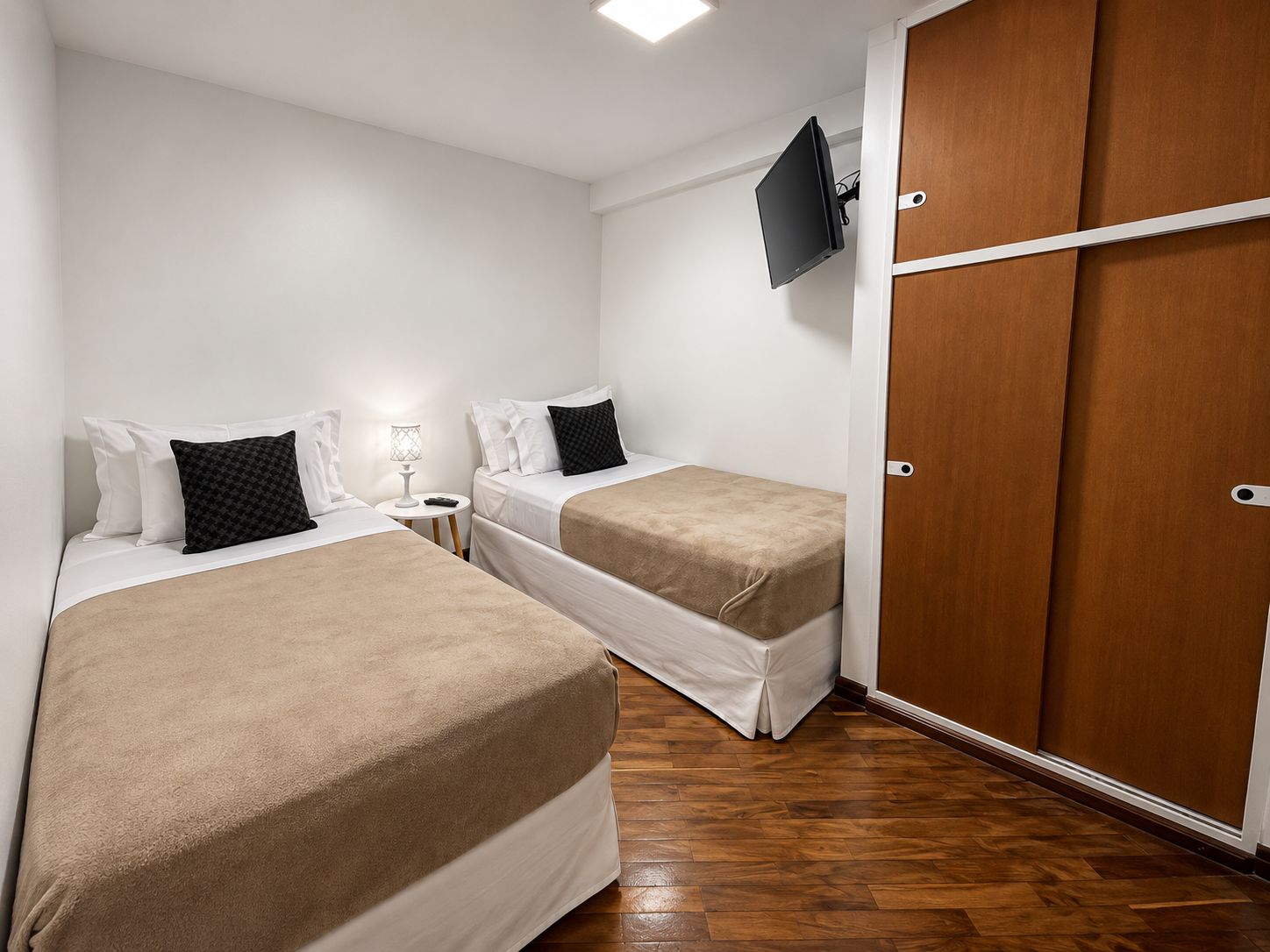Habitación de hotel con dos camas, un televisor montado en la pared, una mesita de noche con lámpara y un armario con espejo.