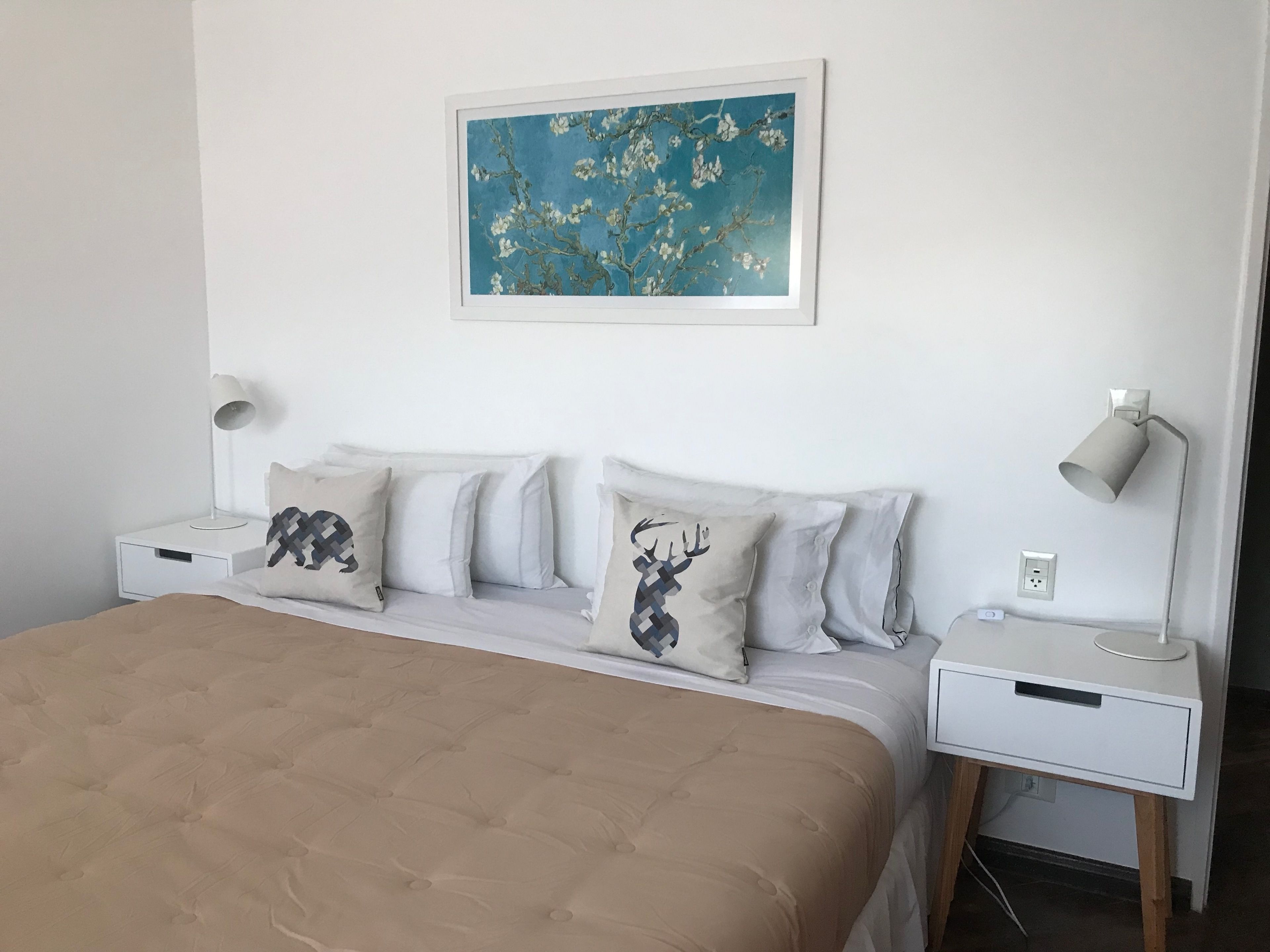 Un dormitorio ordenado con una cama grande, dos mesitas de noche con lámparas y una obra de arte enmarcada en la pared.